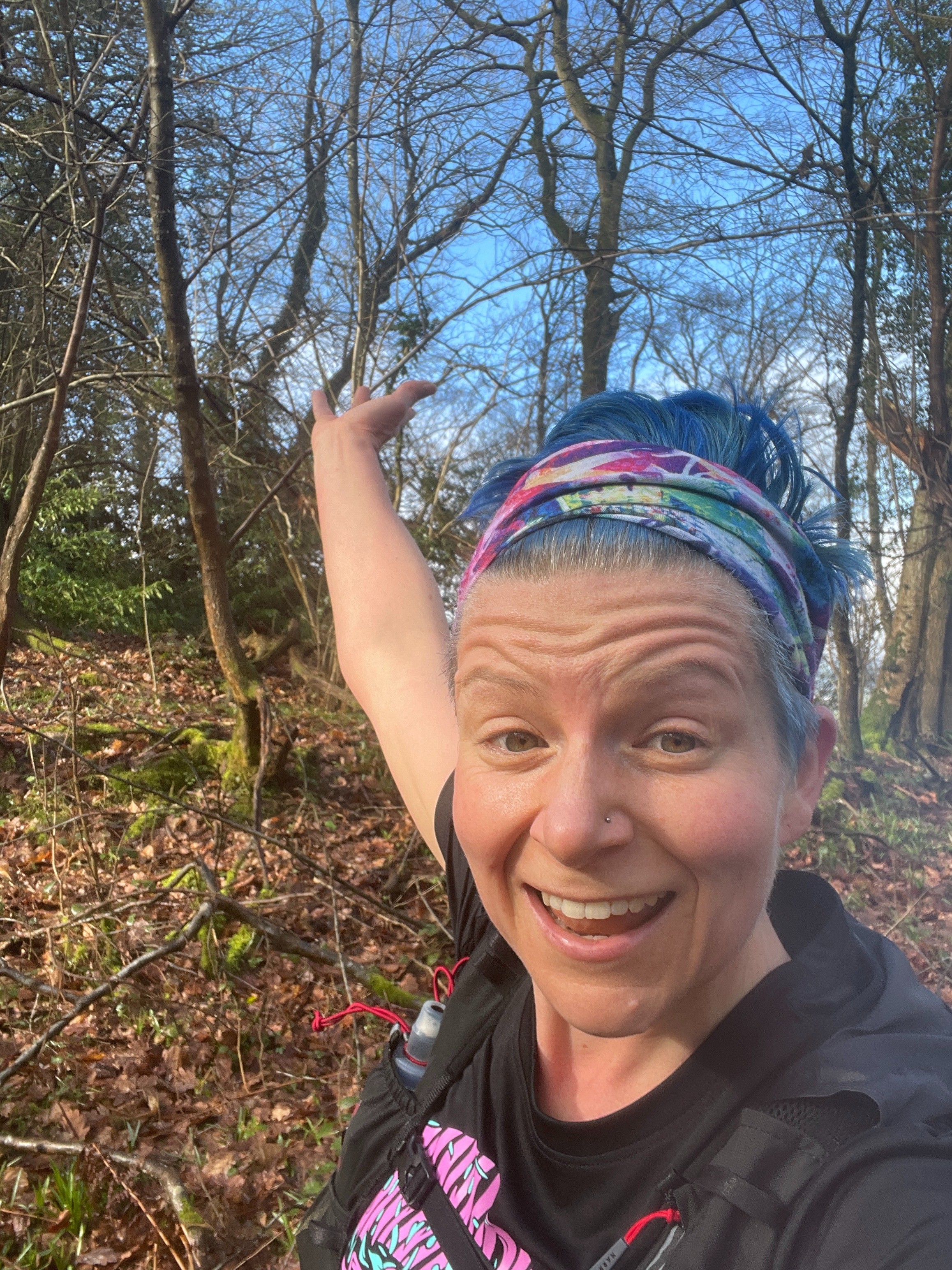 Image shows the head and shoulders of Charlotte, a woman in her mid 40s, on a run in the woods in late winter early spring. Charlotte has short blue hair. She is wearing a black t-shirt. Charlotte is looking at the camera and holding her right arm up, gesturing at her surroundings in a "wow this is amazing" type way.